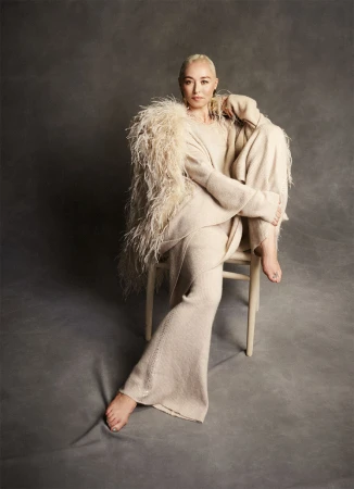 An adult woman with a shaved head poses seated on a chair in a neutral-colored sweater and trousers, featuring a feathered garment on her shoulders.